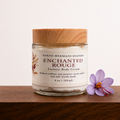 Enchanted Rouge Luxury Body Cream in a clear 4 oz jar with a wooden lid, displayed on walnut beside a purple saffron crocus.
