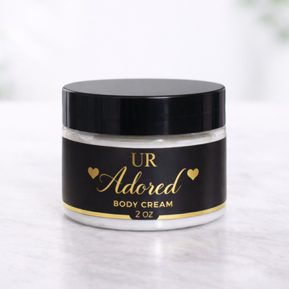 UR Adored 2 oz hydrating body cream made with shea butter, mango butter, jojoba oil, and hyaluronic acid, packaged in a clear jar with black lid and gold foil label on a white marble background.