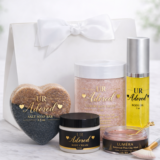 UR Adored Valentine’s Day luxury gift set displayed in a white satin-bow gift bag with a heart-shaped salt soap bar, 7 oz mineral bath salts, 2 oz body oil, 2 oz body cream, and Luméra botanical pink clay face mask with gold spoon and fan brush on a marble background.