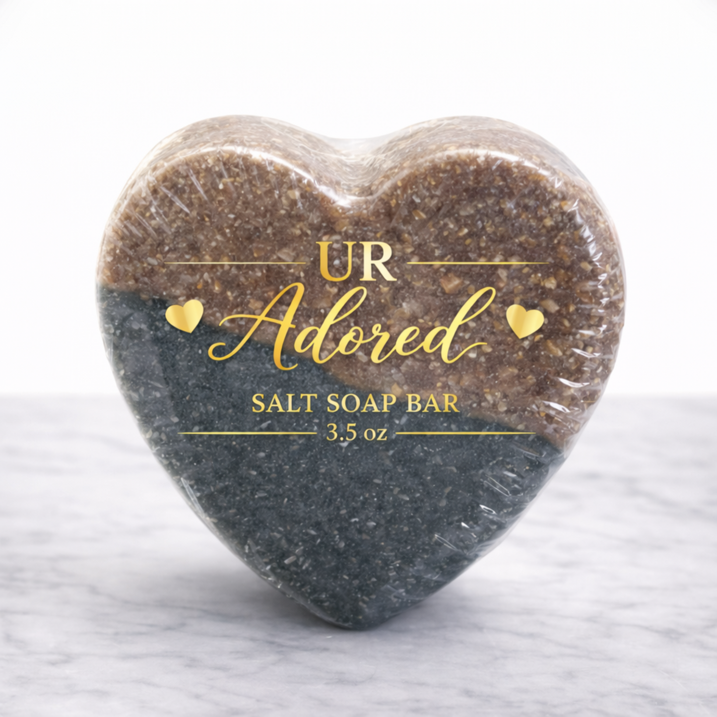 Heart-shaped UR Adored salt soap bar wrapped in clear bio-shrink wrap with gold foil label, featuring a two-tone mineral salt soap in warm brown and deep charcoal on a white marble background.