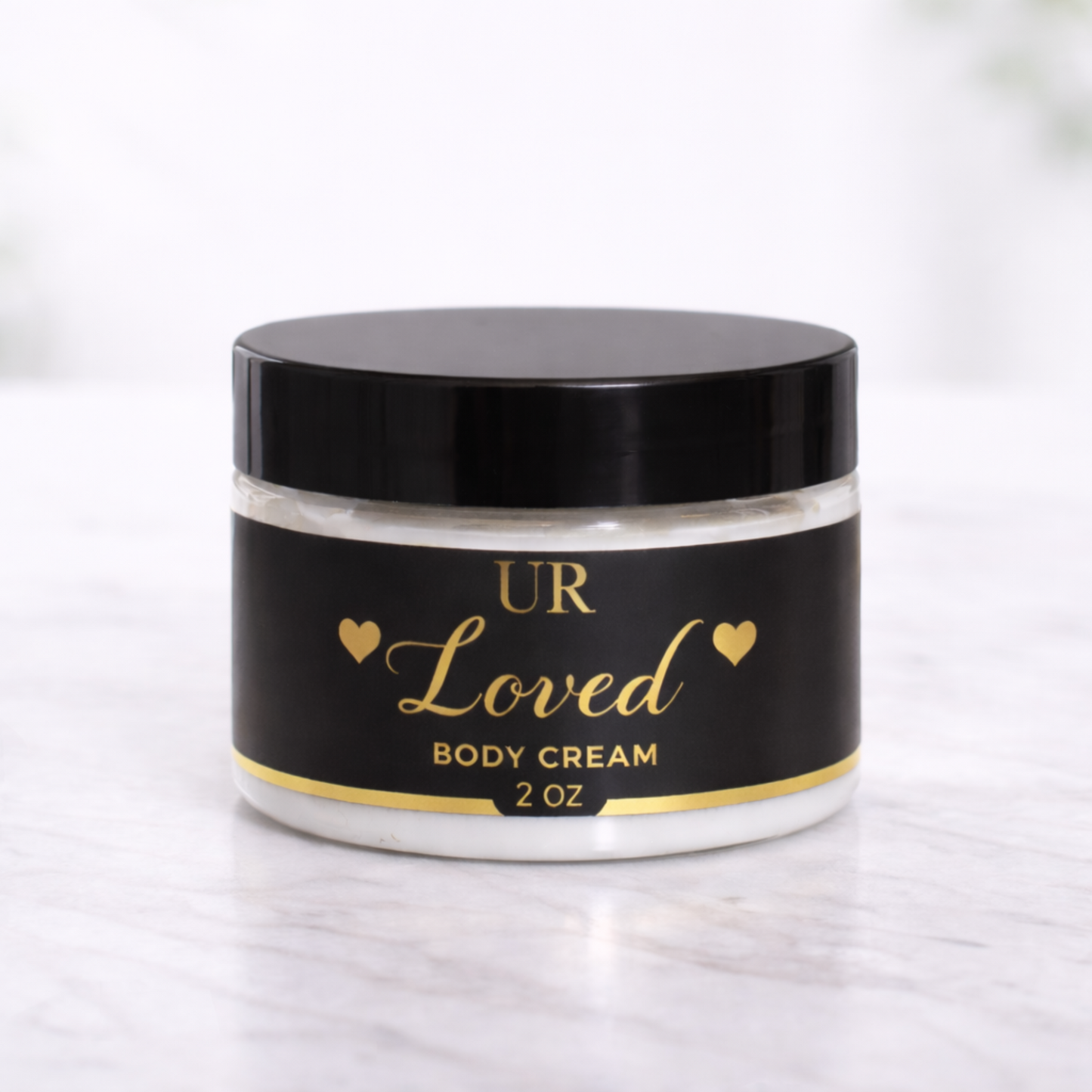 UR Loved 2 oz hydrating body cream made with shea butter, mango butter, jojoba oil, and hyaluronic acid, packaged in a clear jar with black lid and gold foil label on a white marble background.