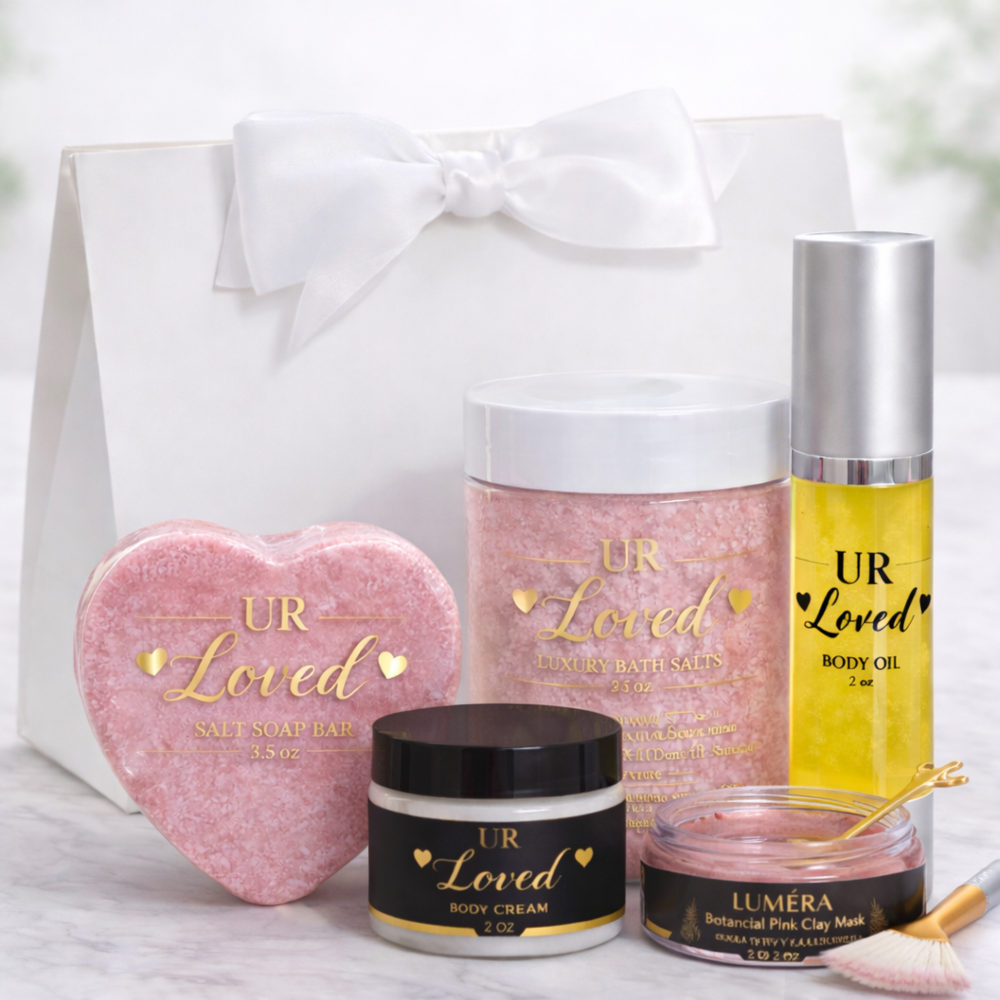“UR Loved Valentine’s Day gift set displayed in a white satin-bow gift bag with a heart-shaped pink Himalayan salt soap bar, luxury mineral bath salts, 2 oz body oil, 2 oz body cream, and Luméra botanical pink clay face mask with gold spoon and fan brush, styled on a marble background.
