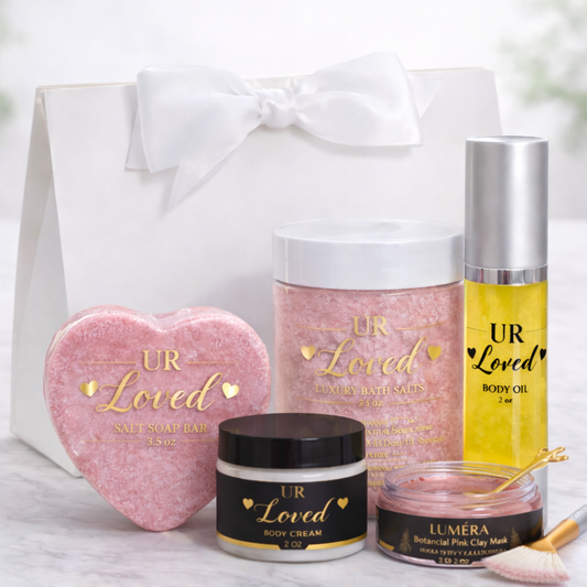 “UR Loved Valentine’s Day gift set displayed in a white satin-bow gift bag with a heart-shaped pink Himalayan salt soap bar, luxury mineral bath salts, 2 oz body oil, 2 oz body cream, and Luméra botanical pink clay face mask with gold spoon and fan brush, styled on a marble background.