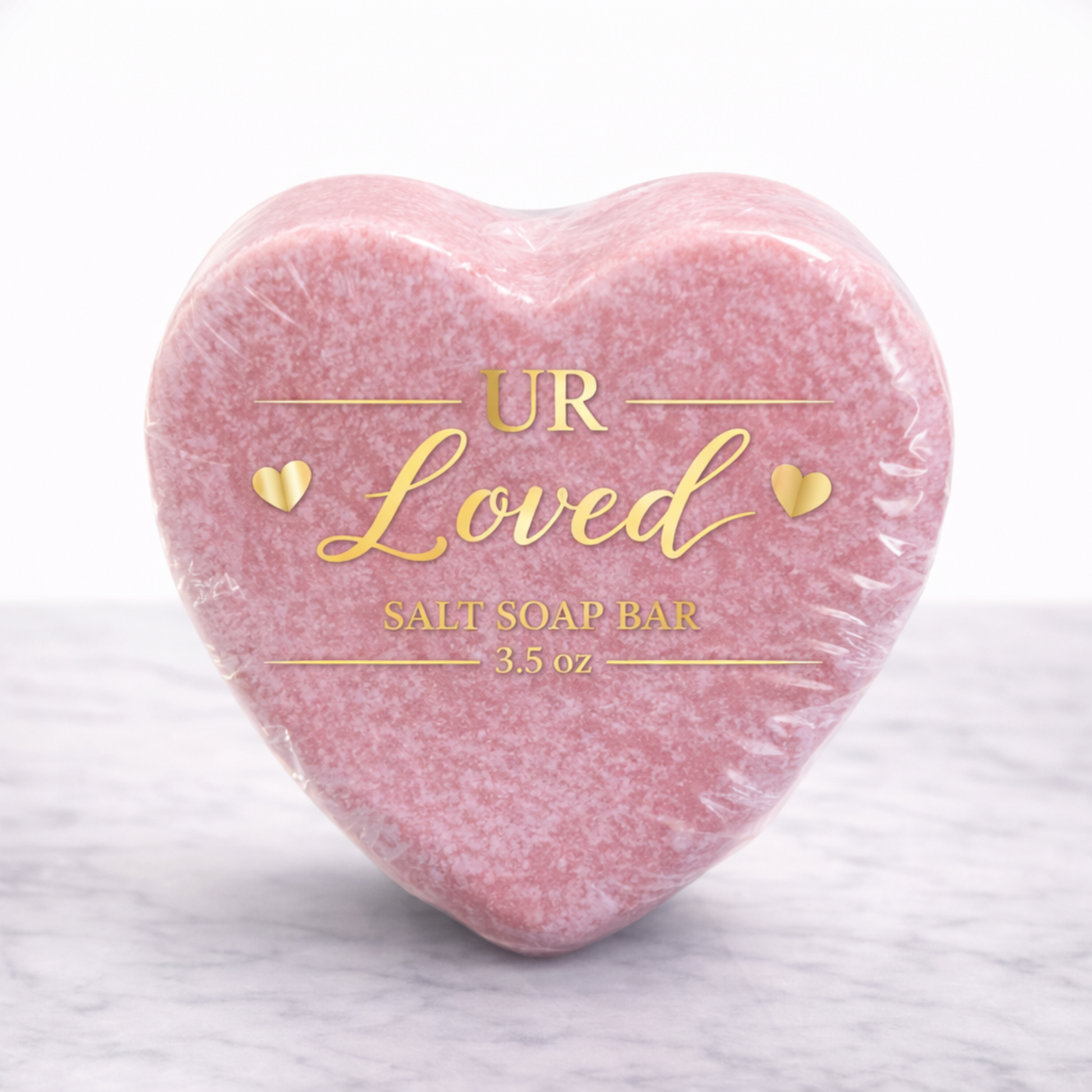 Heart-shaped UR Loved salt soap bar wrapped in clear bio-shrink wrap with gold foil label, featuring soft pink mineral salt soap on a white marble background.