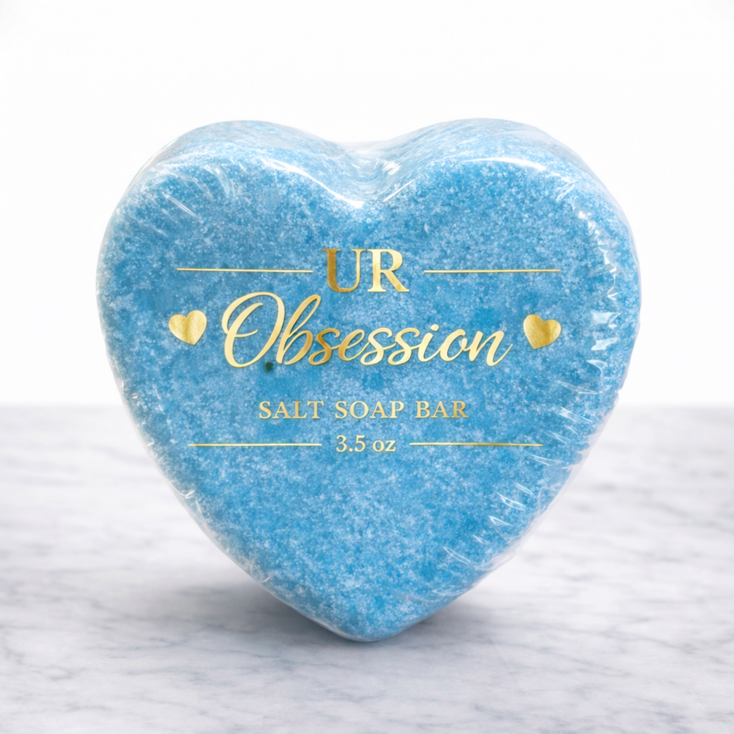Heart-shaped UR Obsession salt soap bar wrapped in clear bio-shrink wrap with gold foil label, featuring vibrant blue mineral salt soap on a white marble background.