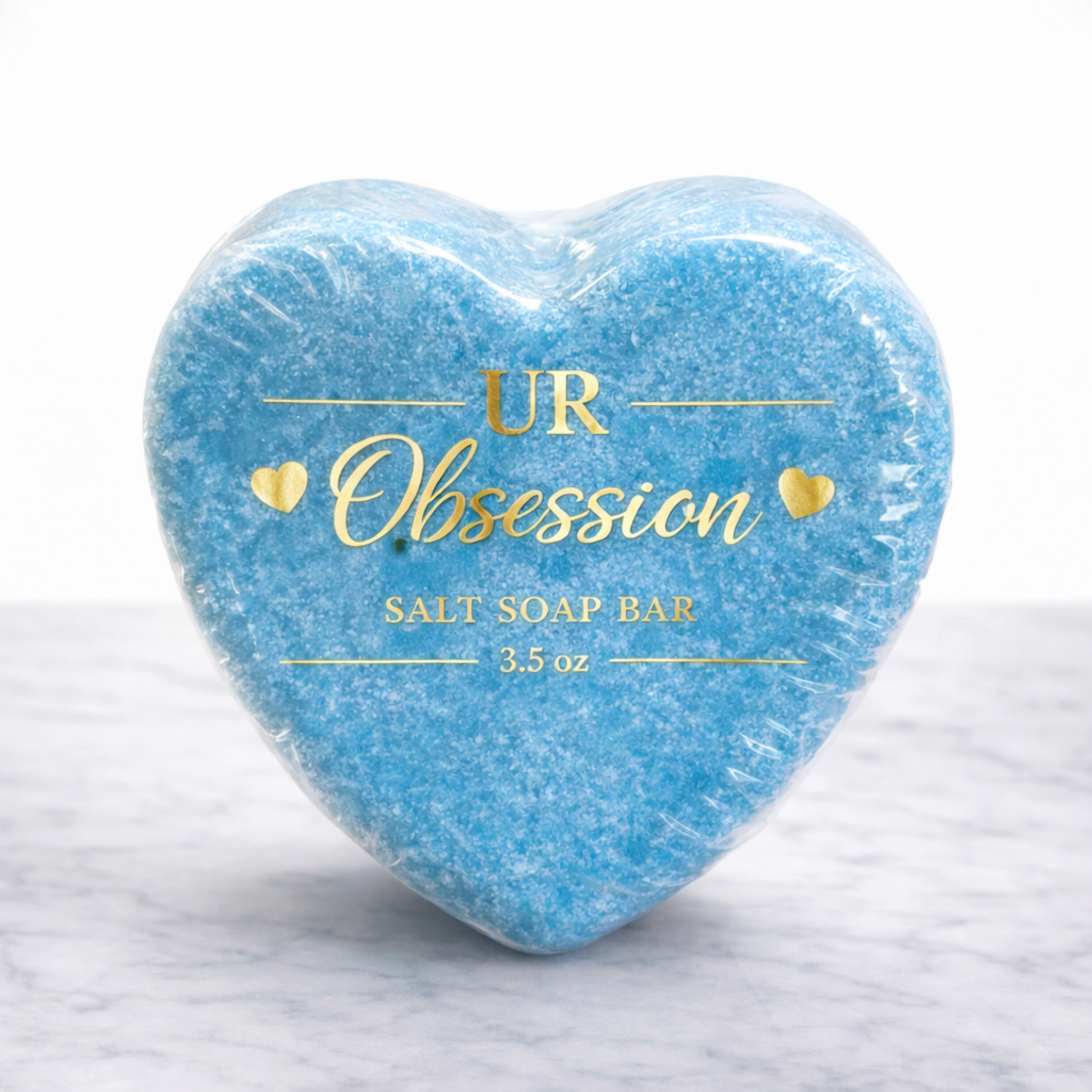 Heart-shaped UR Obsession salt soap bar wrapped in clear bio-shrink wrap with gold foil label, featuring vibrant blue mineral salt soap on a white marble background.