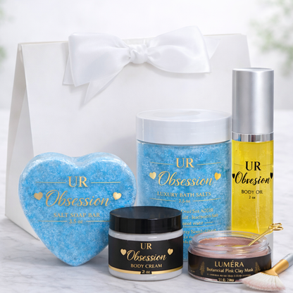 UR Obsession Valentine’s Day luxury gift set displayed in a white satin-bow gift bag with a heart-shaped blue salt soap bar, 7 oz mineral bath salts, 2 oz body oil, 2 oz body cream, and Luméra botanical pink clay face mask with gold spoon and fan brush on a marble background