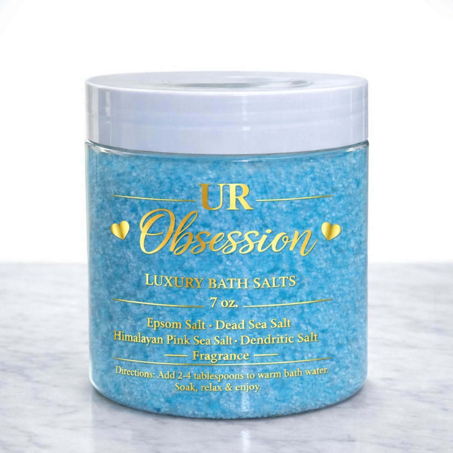 UR Obsession luxury bath salts in a clear 7 oz jar with gold foil label, filled with vibrant blue mineral bath crystals styled on a white marble countertop.
