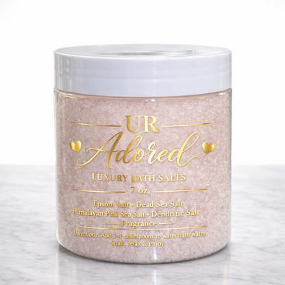 Luxury UR Adored bath salts in a clear 7 oz jar with gold foil label, filled with soft pink Himalayan and Dead Sea salt crystals on a white marble background.
